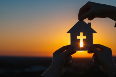 Hands holding house model with cross against sunset sky